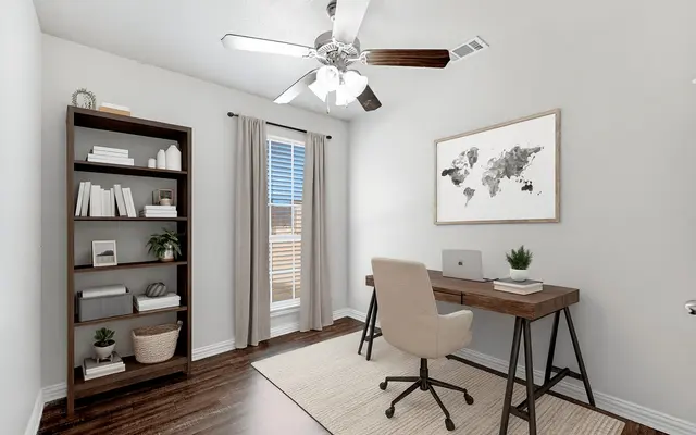 A modern home office featuring a wooden desk with a computer, a comfortable chair, and a wall-mounted world map. A tall bookshelf filled with books and decorative items is on the left side, and light curtains cover a window that lets in natural light. The floor is a warm wood tone, and a beige area rug is underneath the desk.