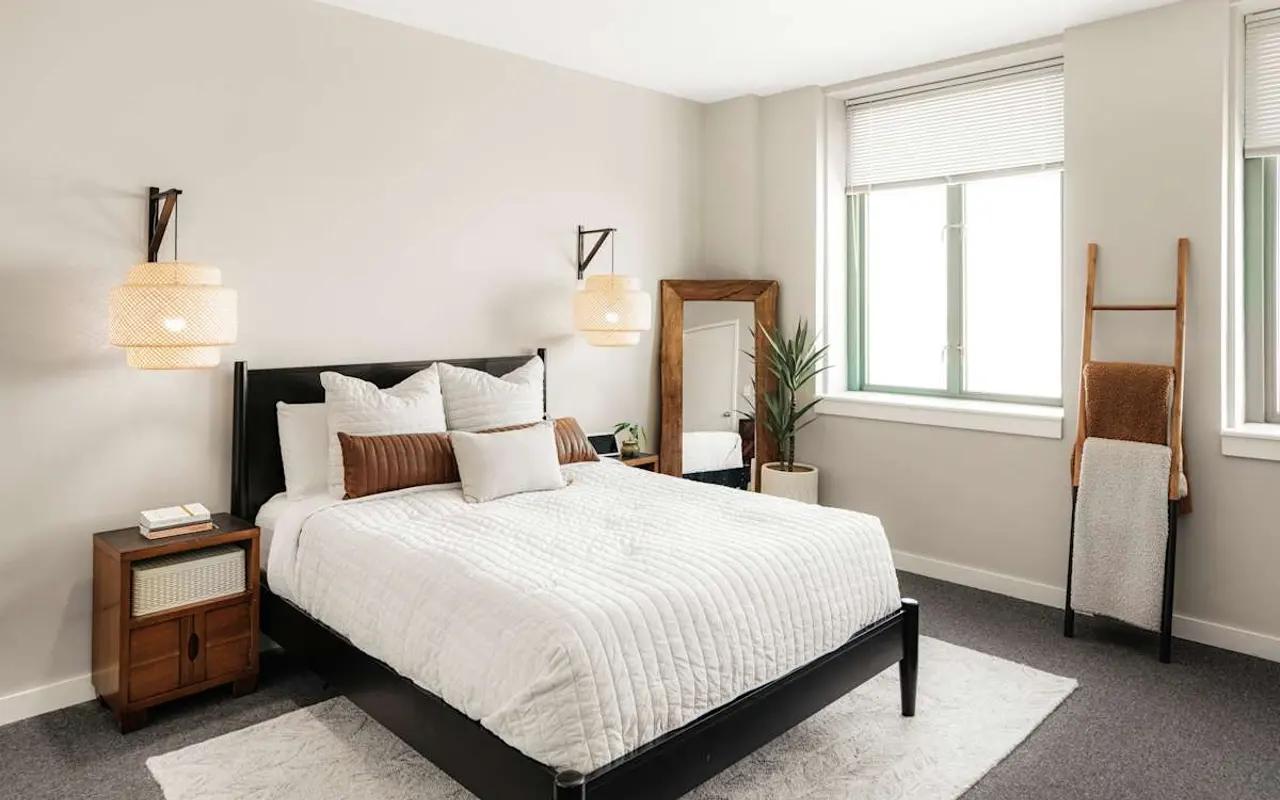 A modern bedroom featuring a black bed with a white quilt, two bedside lamps, a wooden mirror, and a ladder-style shelf against the wall. Large windows allow natural light to enter the room, highlighting the plants and minimalist decor.