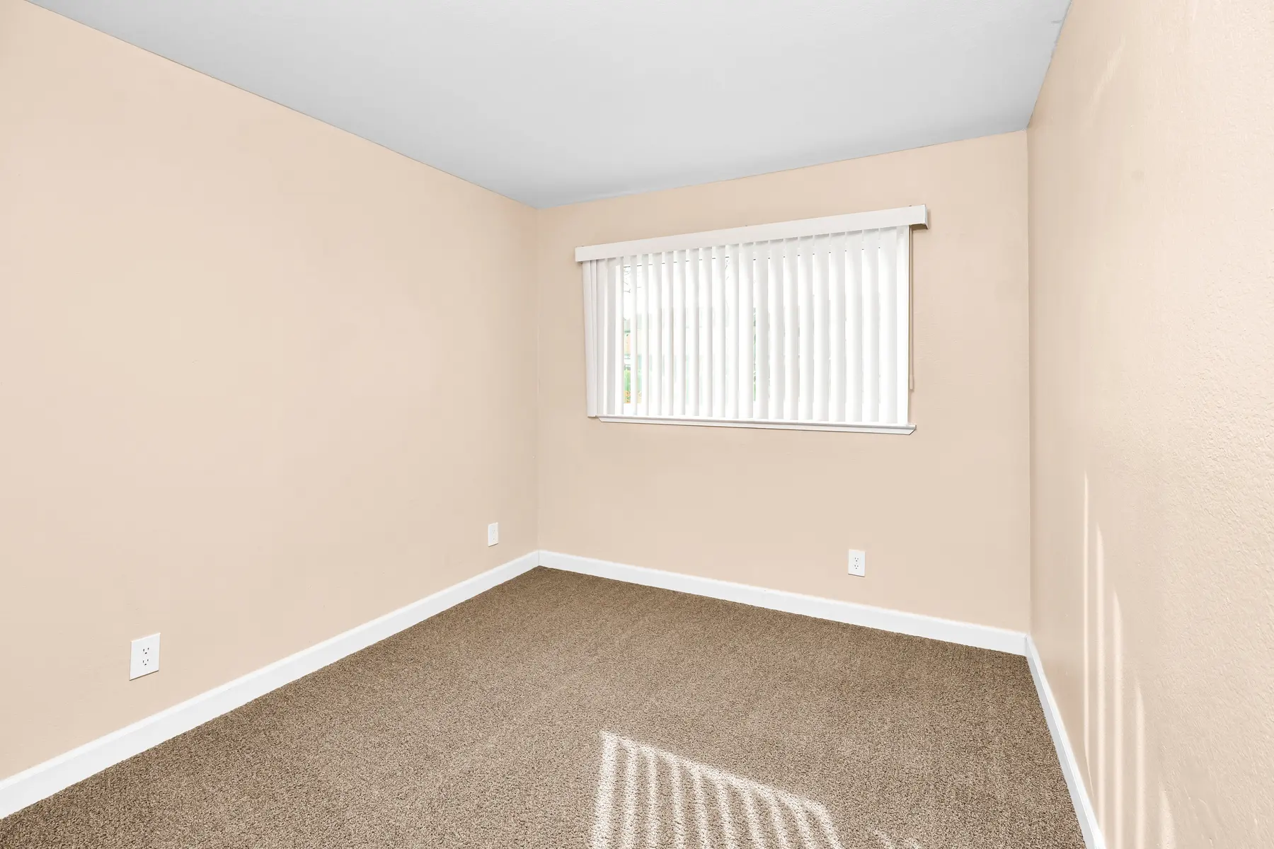 A vacant room with beige walls and a light brown carpet. A window with vertical blinds allows natural light to enter the space.