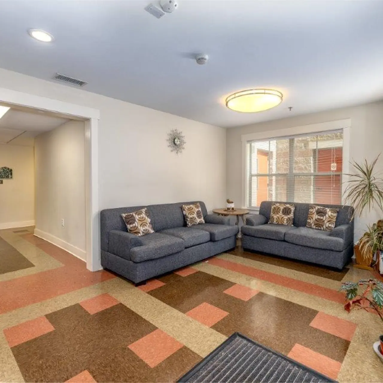A cozy lobby area featuring two gray sofas adorned with decorative pillows, tiled flooring in various colors, potted plants, and a wall clock. The entrance door is visible leading to another area.