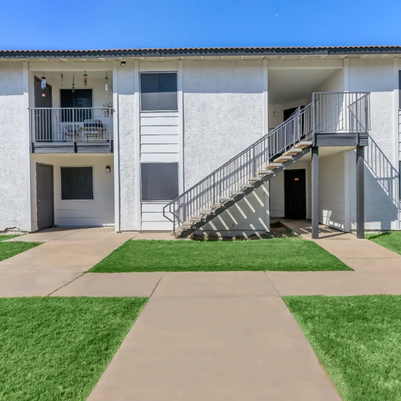 Exterior view of a two-story apartment building with a staircase and a pathway in front, surrounded by grassy areas.