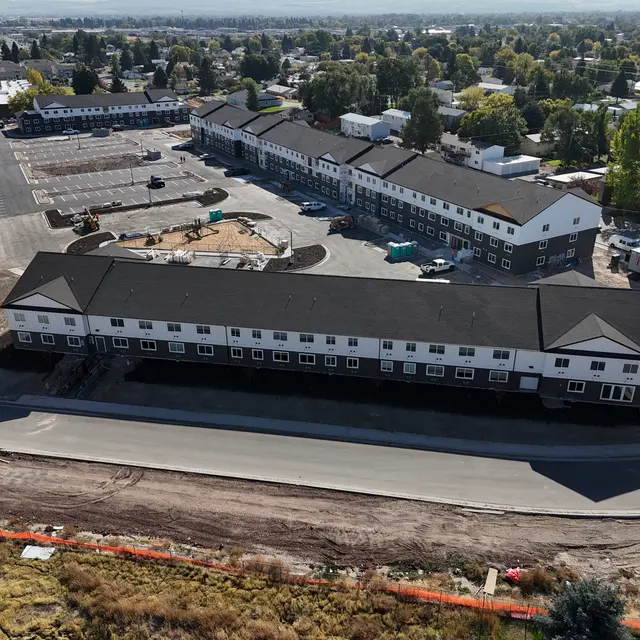 Aerial View of Construction Site Aerial view of a construction site featuring a new building and parking area. The area includes several buildings with uniform architecture, parked vehicles, and surrounding trees.