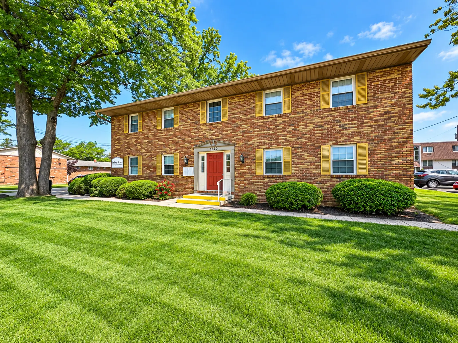 Exterior view of a brick apartment building with yellow shutters and a red door, surrounded by green grass and trees.