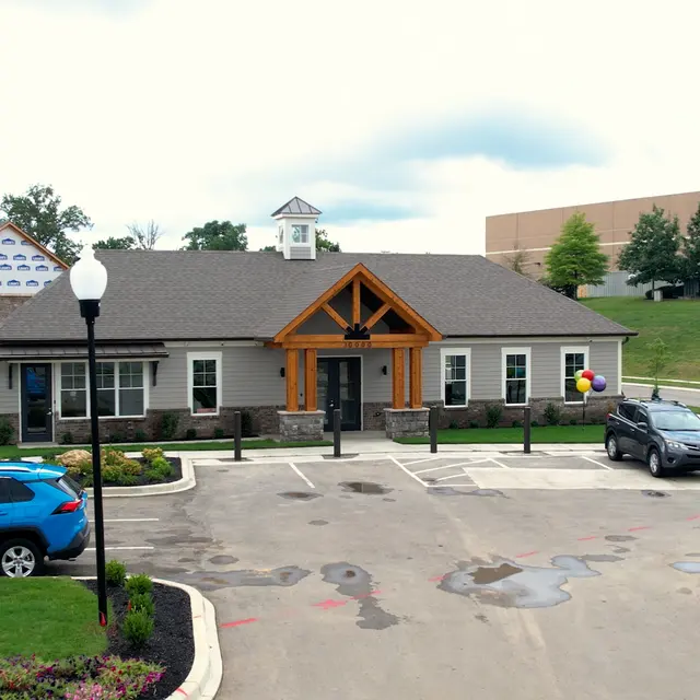 A modern building with a stone and wood exterior, located in a parking lot with cars. There is a decorative sign and some greenery in the surroundings.