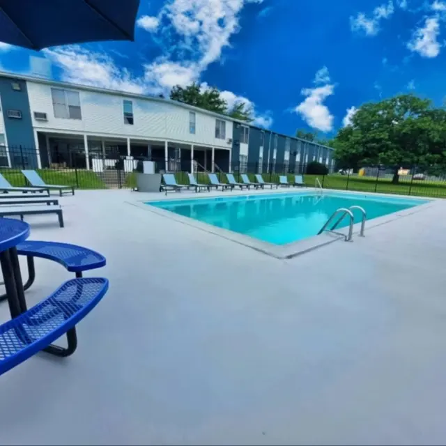 A brightly lit pool area featuring blue lounge chairs, tables, and a clear sky in the background.