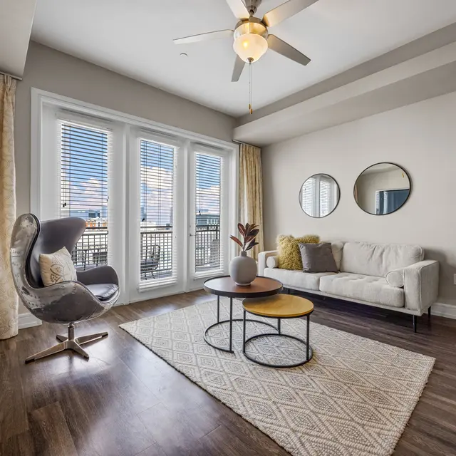 A modern living room featuring a light-colored sofa, a gray accent chair, a round coffee table with two tiers, and a decorative plant. Large windows with white blinds allow natural light to enter, while walls are painted in soft tones. Two round mirrors adorn the wall opposite the sofa.