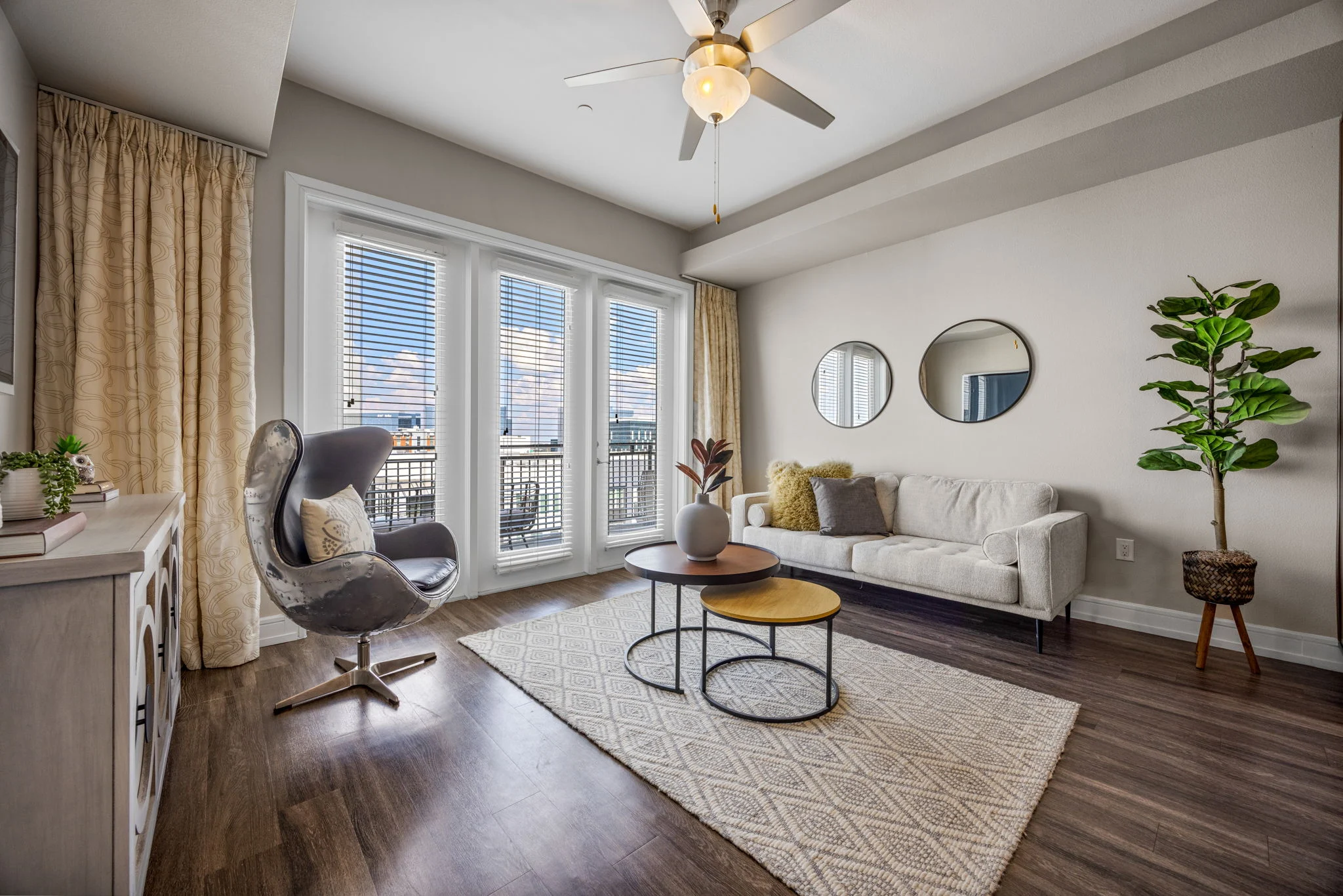 A modern living room featuring a light-colored sofa, a gray accent chair, a round coffee table with two tiers, and a decorative plant. Large windows with white blinds allow natural light to enter, while walls are painted in soft tones. Two round mirrors adorn the wall opposite the sofa.