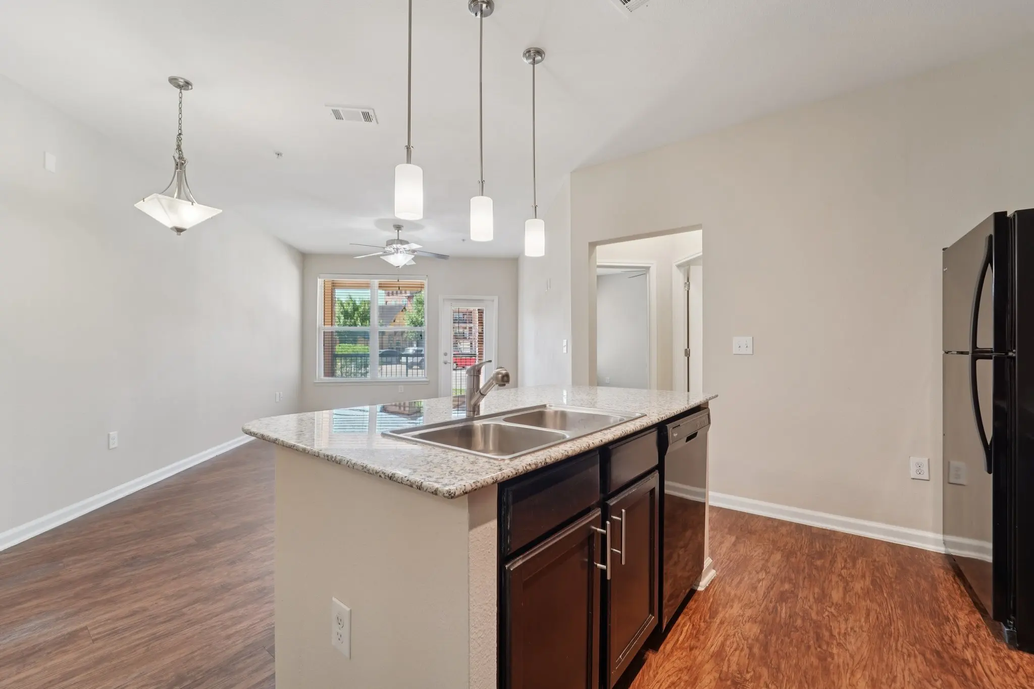 A modern kitchen featuring dark cabinetry, stainless steel appliances, and a granite countertop with a sink. Pendant lights hang above an island, with an open layout leading to a living area.