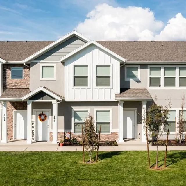 A row of modern townhouses with a well-manicured lawn and decorative features. Each unit has a small porch and seasonal decorations. The sky is partly cloudy.
