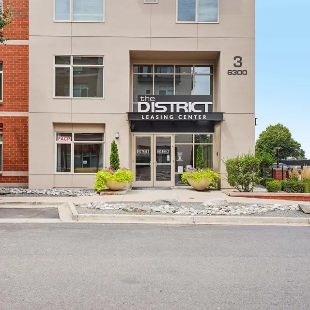 The District Leasing Center Entrance The exterior of a modern building featuring the sign 'the DISTRICT LEASING CENTER' with large windows and planters in front.