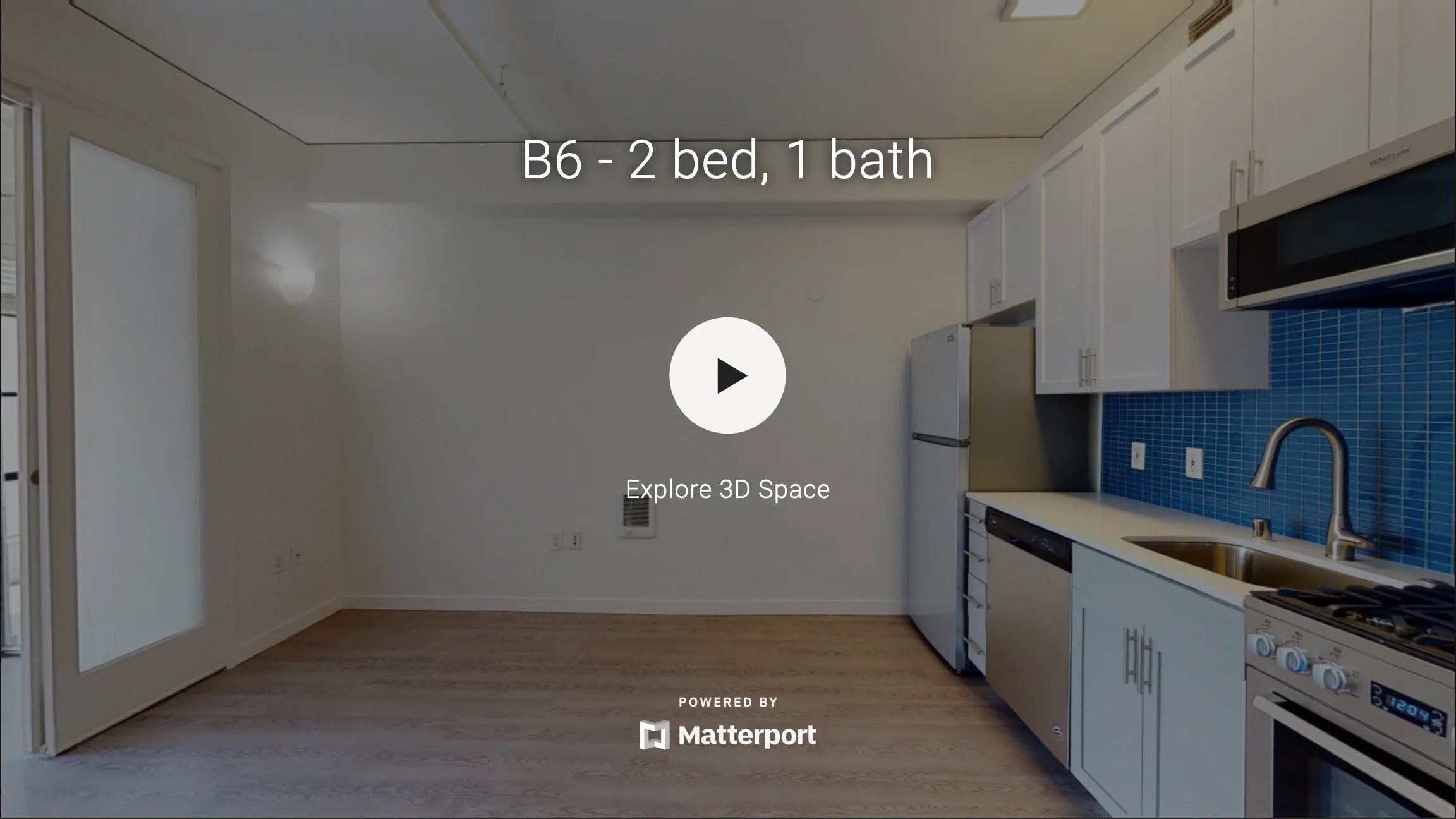 Interior view of a modern apartment showing a kitchen and living area, featuring white cabinets and blue tiled backsplash.