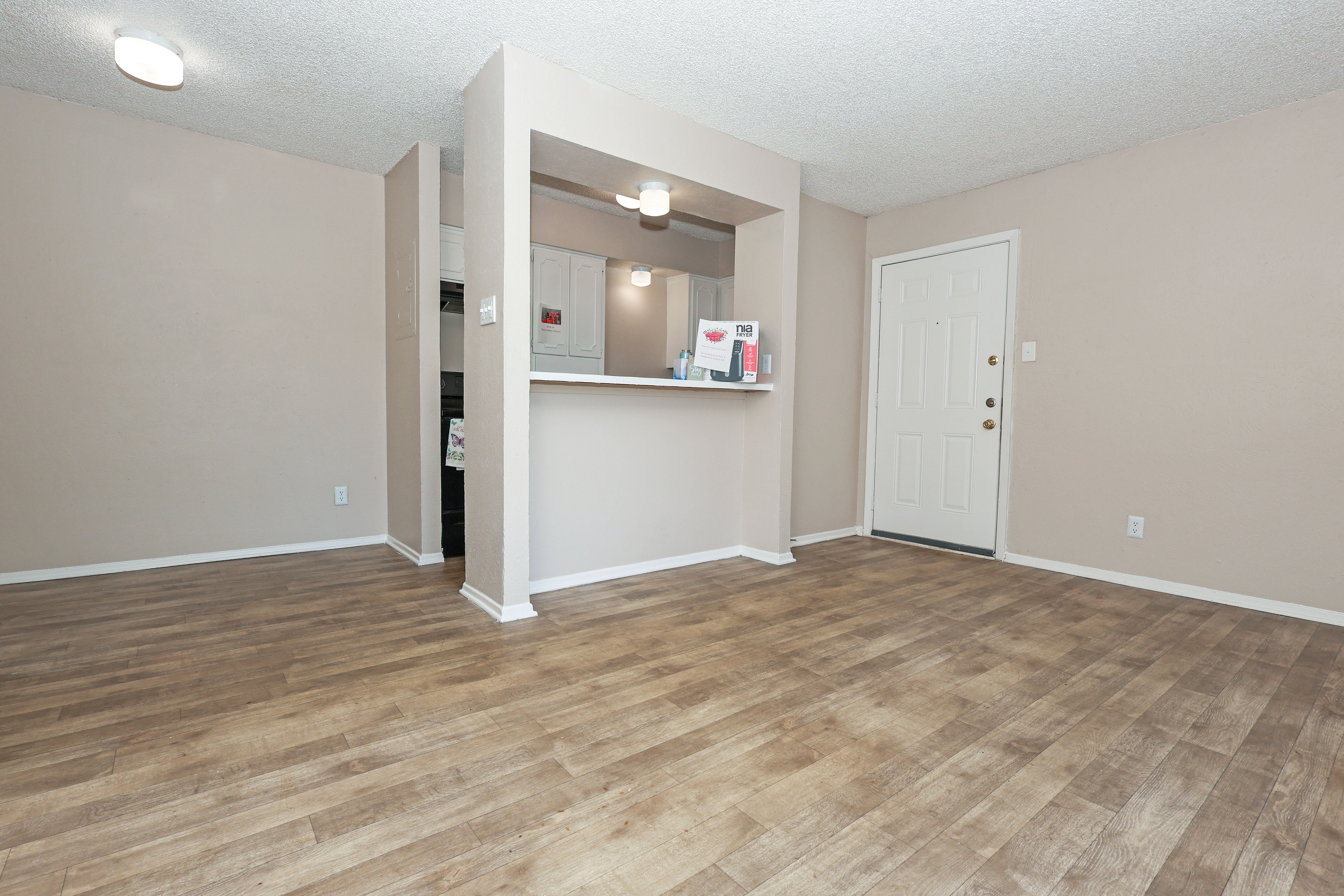 A spacious, empty living room with wooden flooring, beige walls, and a view of an entry door and a small kitchen area.