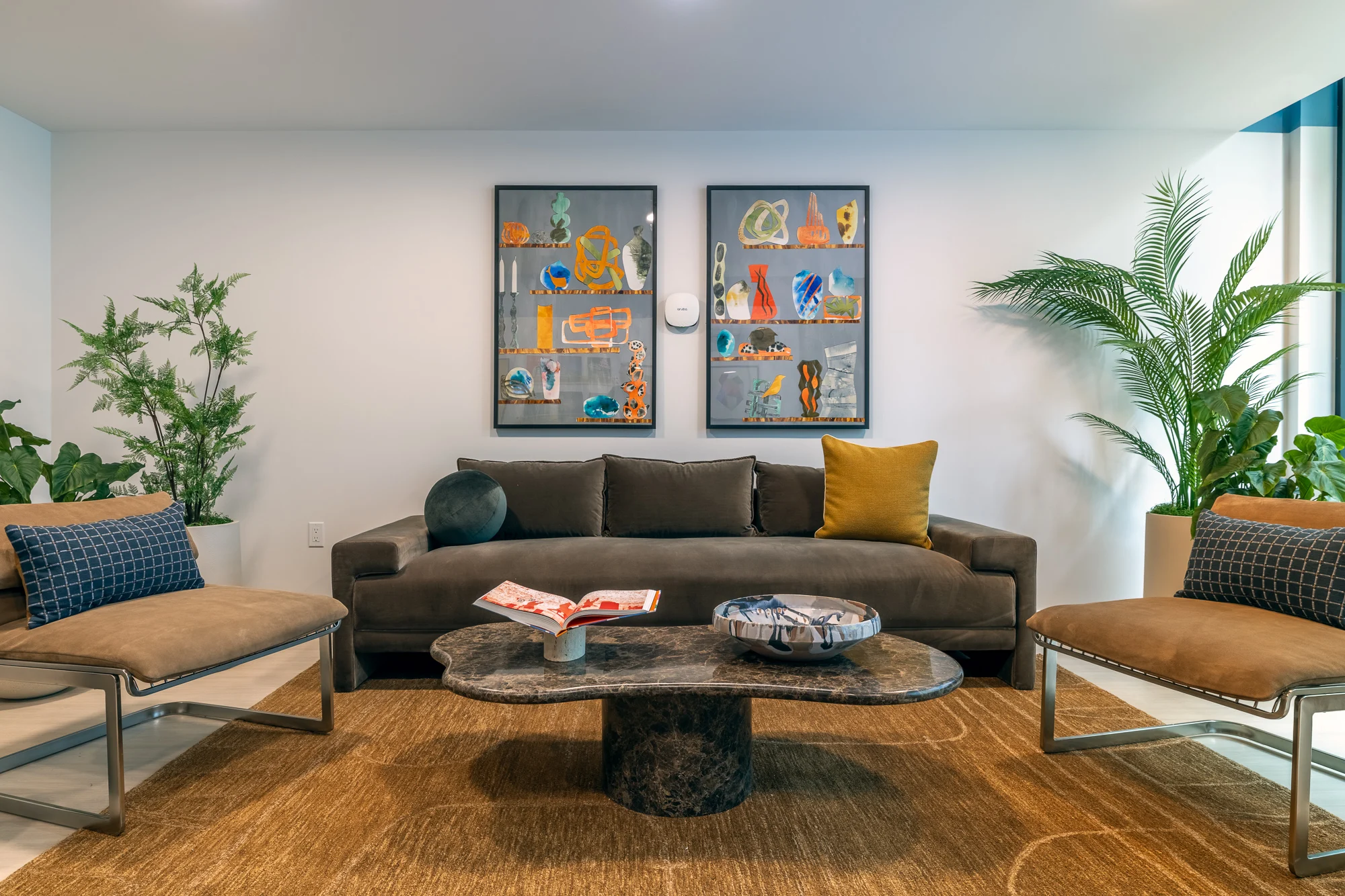 Modern Living Room Design A modern living room featuring a dark sofa with colorful cushions, two stylish chairs, a round coffee table with decorative items, and vibrant artwork on the walls, surrounded by indoor plants.