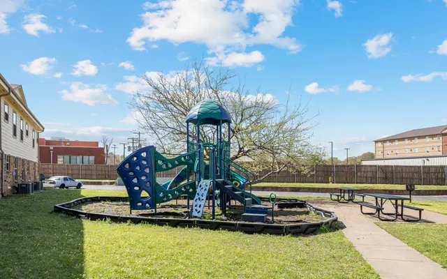 Community Playground A playground structure with blue and green components in a grassy area, surrounded by concrete walkways and a picnic table.