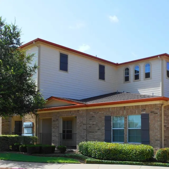 Residential Building Exterior A two-story residential building with a combination of brick and siding exterior, featuring a sloped roof with orange accents and a stone chimney. Lush green bushes and trees line the front of the property under a clear blue sky.