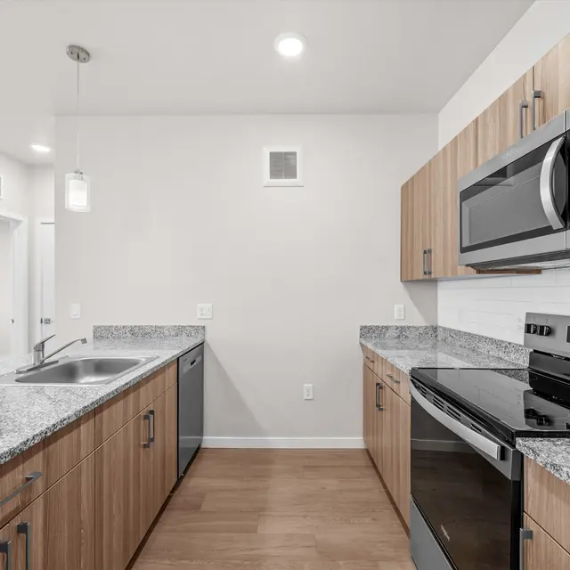 A modern kitchen with wooden cabinets, granite countertops, and stainless steel appliances. It features a clear countertop area, a sink, and hanging lights above.