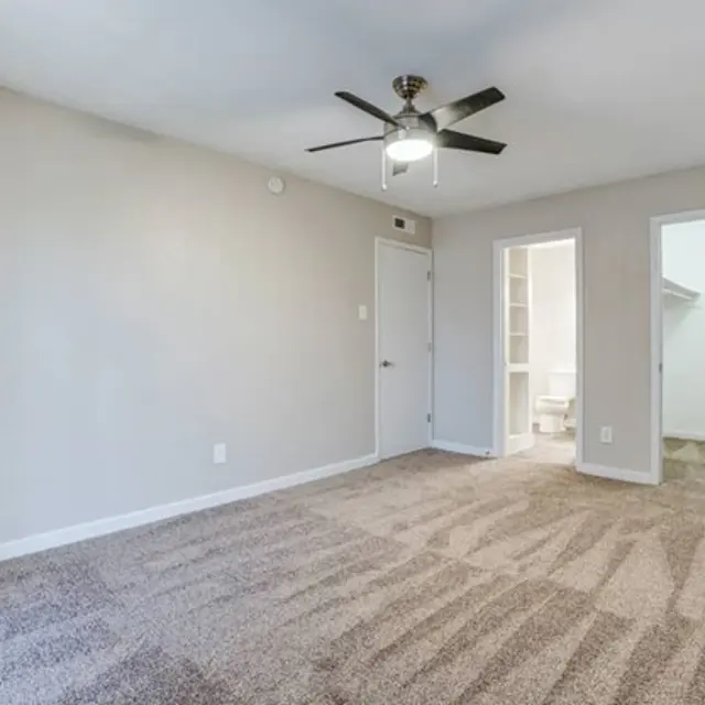 An empty bedroom featuring beige walls, a ceiling fan, and plush carpet flooring. A door leading to a closet is visible, as well as an open door to a bathroom area.