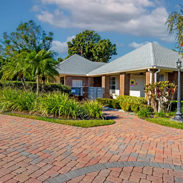 A scenic view of a house with a grey roof, surrounded by lush green plants and a brick driveway, under a blue sky with fluffy clouds.