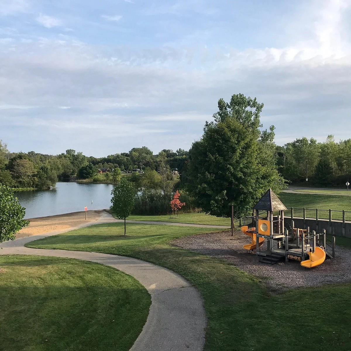 A scenic view of a park featuring a playground with slides, a pathway leading to a small lake, and lush greenery surrounding the area.
