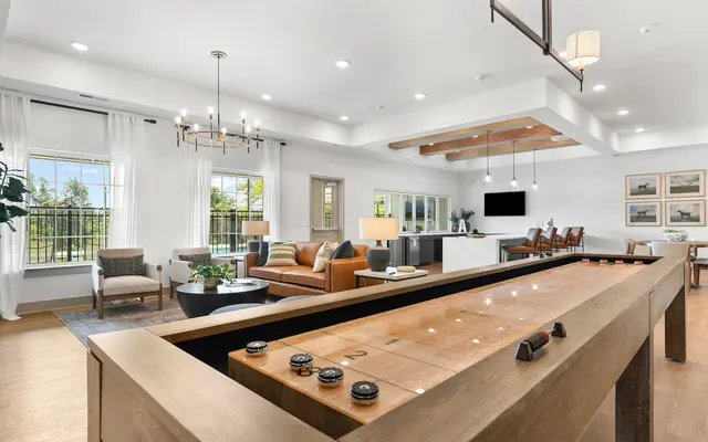 A modern living room featuring a shuffleboard table, a cozy seating area with multiple chairs and a sofa, large windows with white curtains, and a dining area in the background.