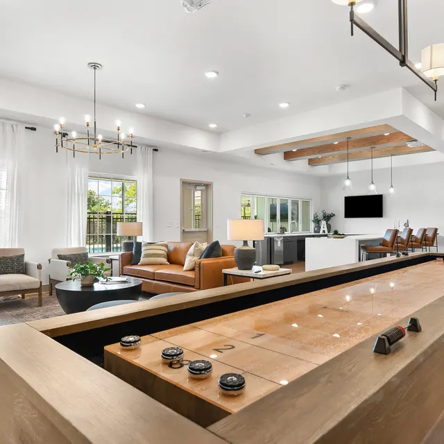 A modern living room featuring a shuffleboard table, a cozy seating area with multiple chairs and a sofa, large windows with white curtains, and a dining area in the background.
