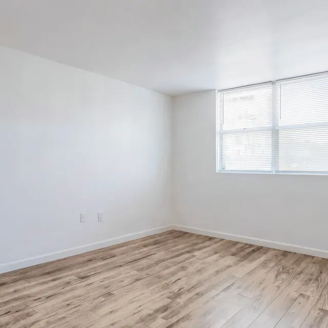 A spacious empty room with light-colored walls and newly installed wooden flooring. A large window allows natural light to fill the space, and a doorway is visible in the background.