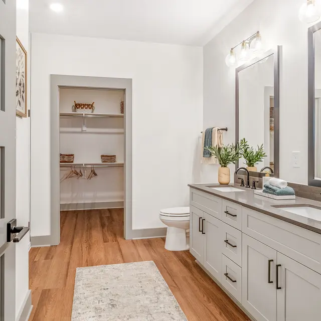 A spacious modern bathroom featuring a double vanity with mirrors, a toilet, and a walk-in closet. The decor is neutral with light wood flooring and greenery accents.