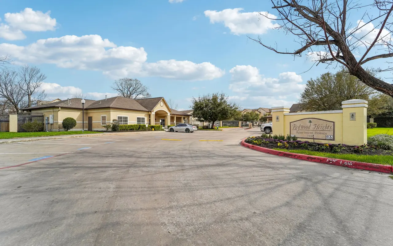 A view of a residential complex entrance with a sign. The background shows a single-story building surrounded by trees and a cloudy blue sky.