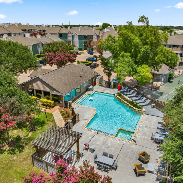Aerial view of a residential complex showcasing a swimming pool surrounded by lounging chairs, with a community building nearby and landscaped trees and flowers.