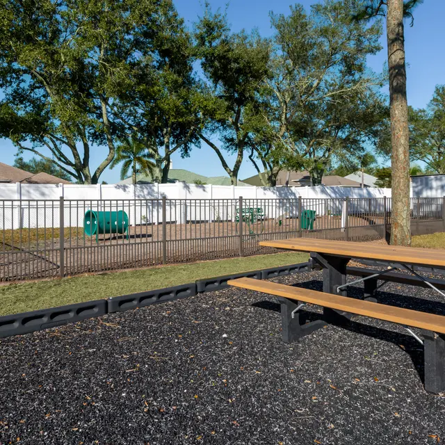 A fenced dog park area with green agility equipment and a picnic table in the foreground.