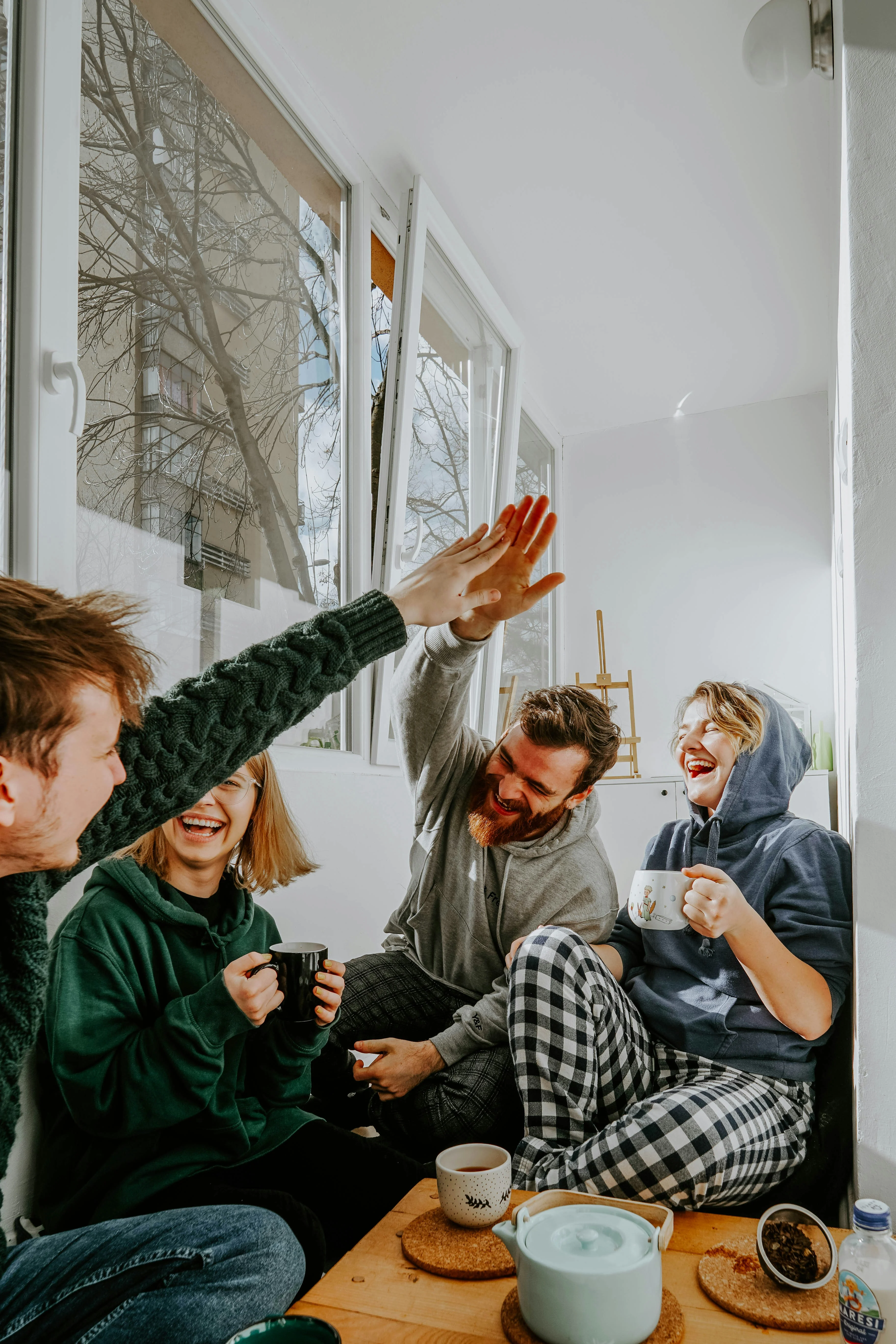 A group of four friends sitting in a cozy corner, laughing and enjoying their time together with mugs in hand. Two are wearing comfortable hoodies and the other two are in relaxed attire. Sunlight streams in through the window.