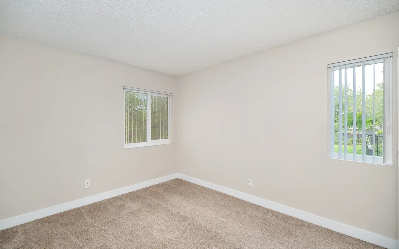 A light-colored empty room with carpeted flooring and two windows with blinds.