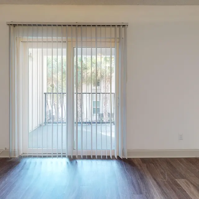 An empty room with wooden flooring and sliding glass doors leading to a balcony, covered with vertical blinds.