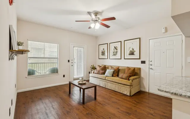 Modern Living Room Interior A bright and inviting living room featuring a beige couch with decorative pillows, a small coffee table in front, and framed botanical art on the wall. Natural light streams in through a window and a door, with a ceiling fan overhead.