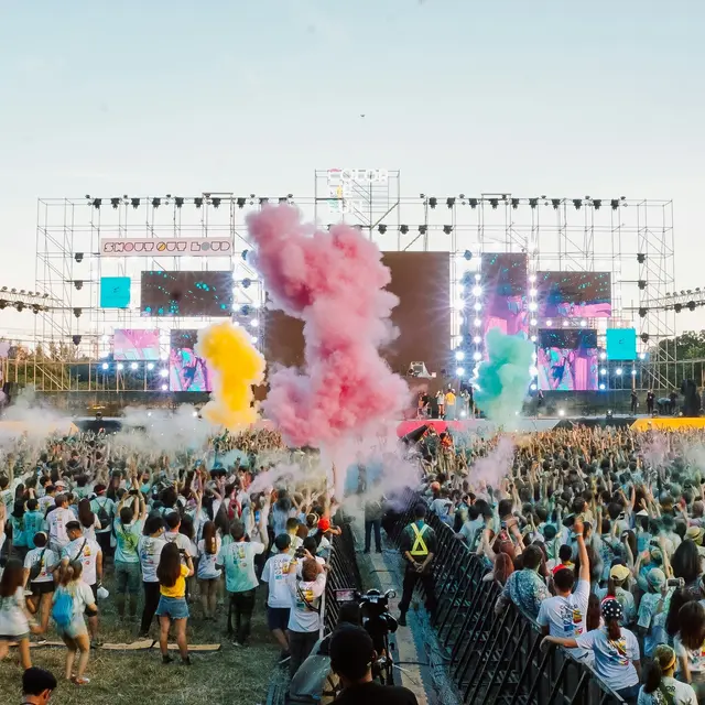 Summerfest A large crowd at a music festival with colorful smoke flares in the air and a vibrant stage in the background.