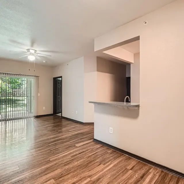 A spacious living room with wood-like flooring, featuring a ceiling fan and large windows with blinds allowing natural light. There is a small breakfast bar connecting to the kitchen area in the background.