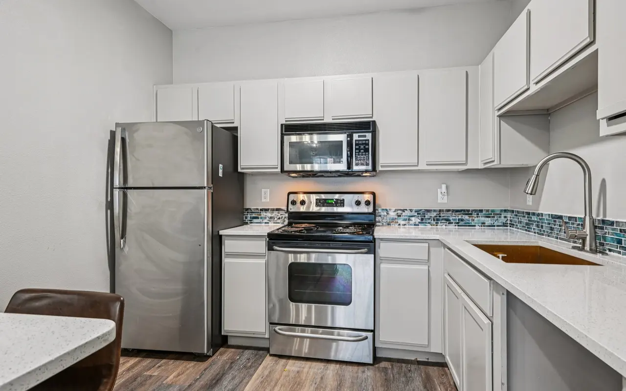 A modern kitchen featuring stainless steel appliances, including a refrigerator, microwave, and stove. The cabinets are white with a blue tile backsplash and a sink.