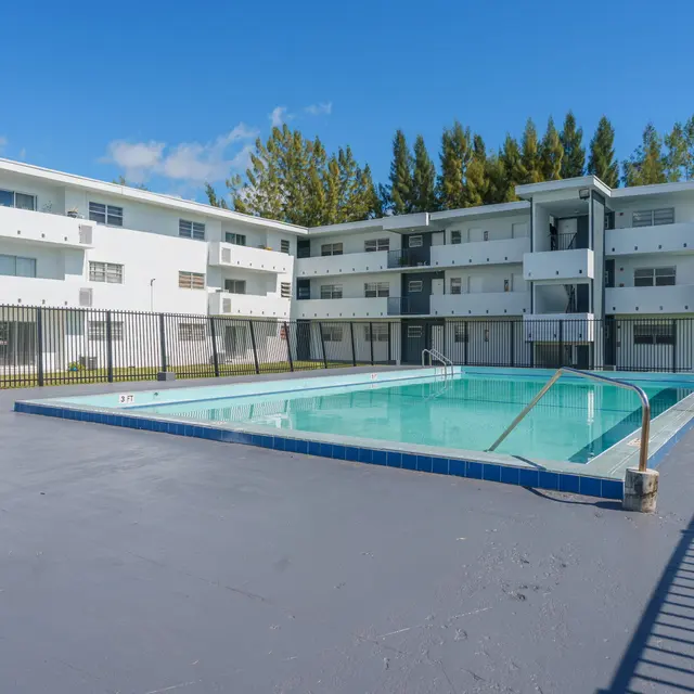 An outdoor swimming pool surrounded by a fence, with an apartment building in the background.