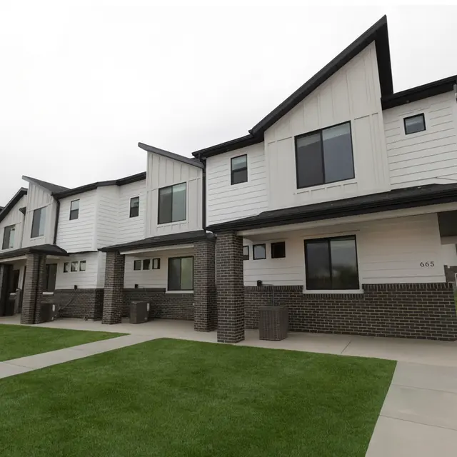 A series of modern townhouses with a laid-out pathway and manicured lawn in front. The buildings have a mix of white and brick exteriors, featuring multiple windows and a clean architectural design.