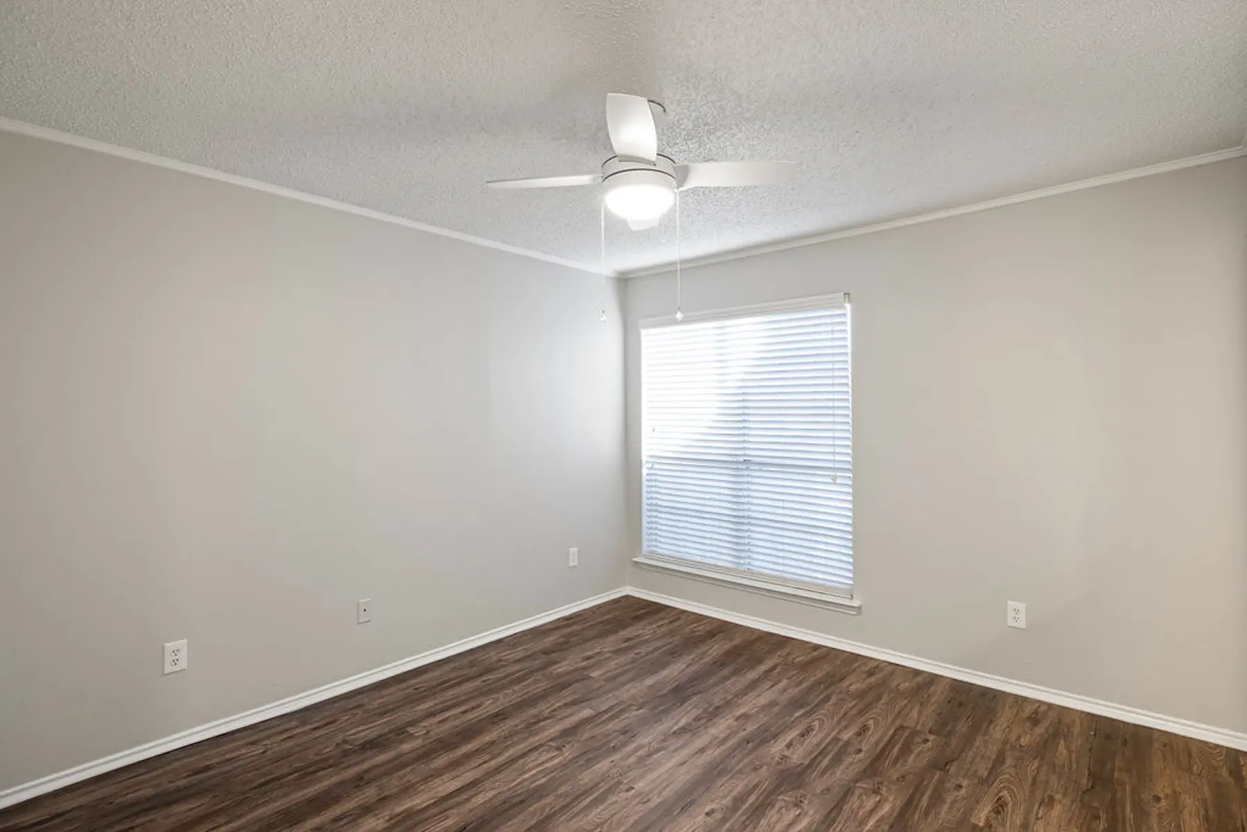 An empty room with wooden flooring, a ceiling fan, and a window with blinds.