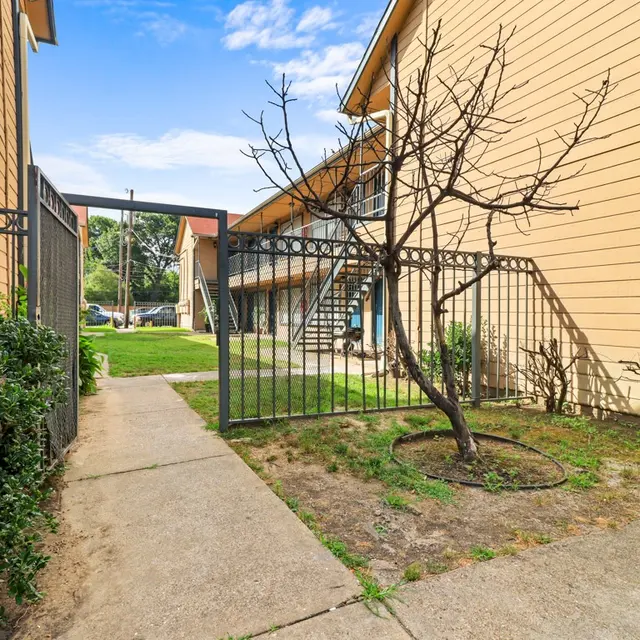 View of a paved pathway leading to a gated entrance, flanked by small green shrubs and a bare tree, with apartment buildings in the background.