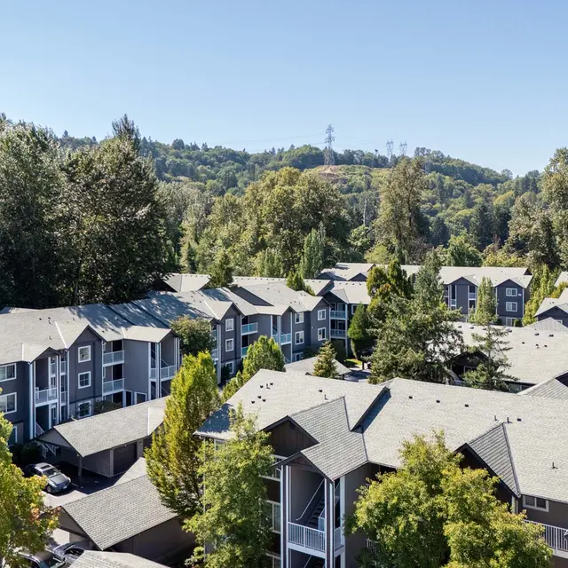 Aerial view of a residential apartment complex surrounded by trees and greenery, with distant hills in the background under a clear blue sky.
