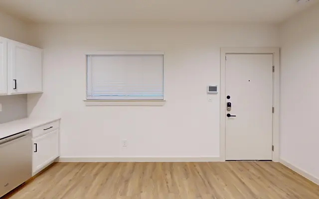 A minimalist, empty room featuring a kitchenette with white cabinets and a window, along with a door on the right side.