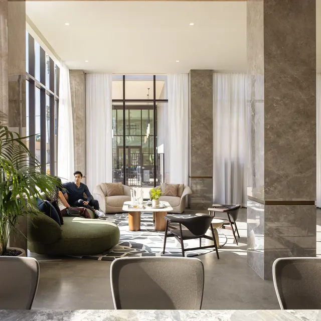 A modern lobby interior featuring large windows, marble columns, and a warm color palette. A person is seated on a sofa with a round coffee table in front, surrounded by greenery and stylish furniture.