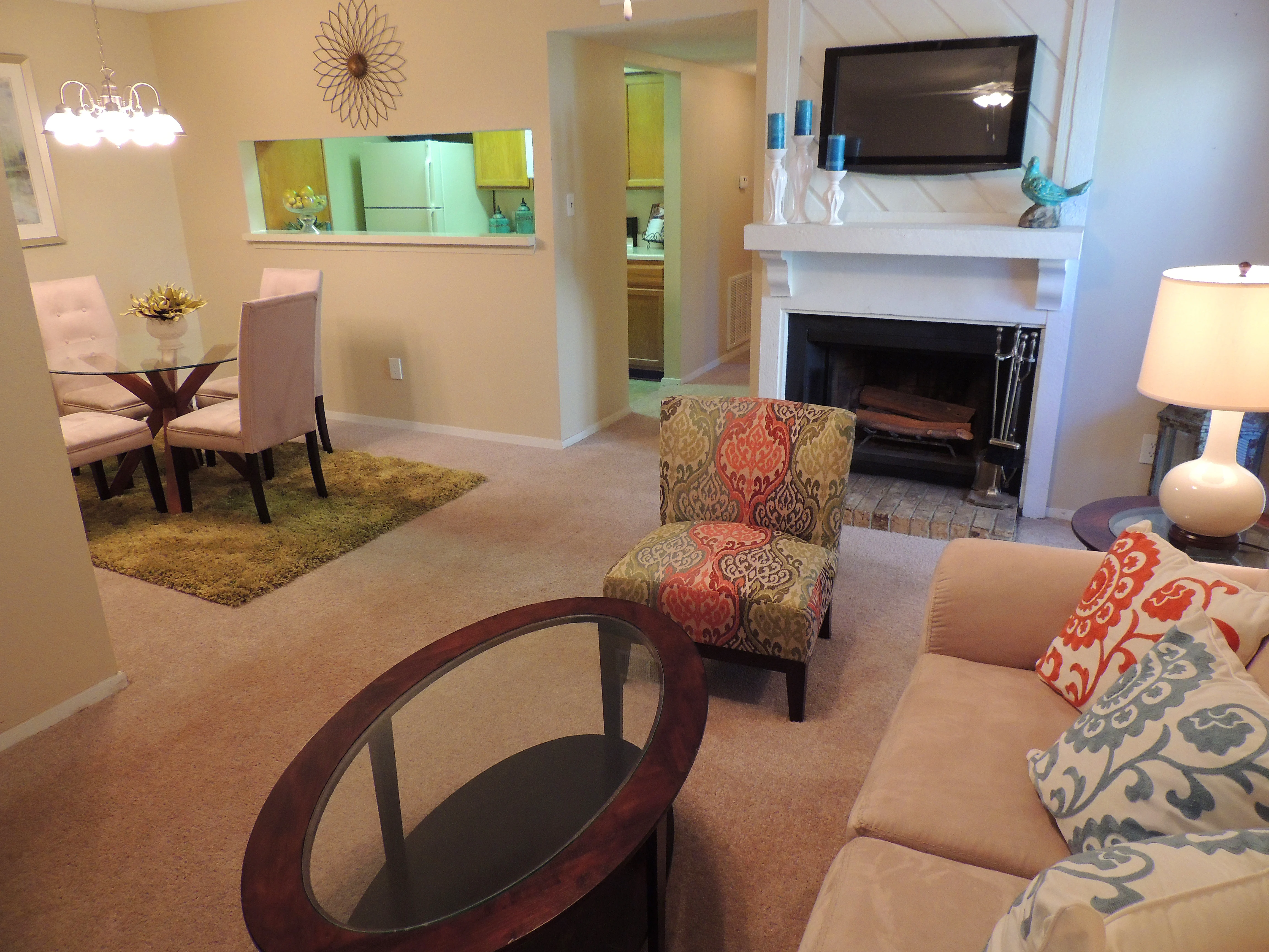 A cozy living room featuring a beige sofa with decorative pillows, a circular wooden coffee table, and an accent chair with a colorful pattern. The space has a fireplace, a TV mounted on the wall, and dining area visible in the background with a round table and chairs. Soft lighting adds warmth to the room.