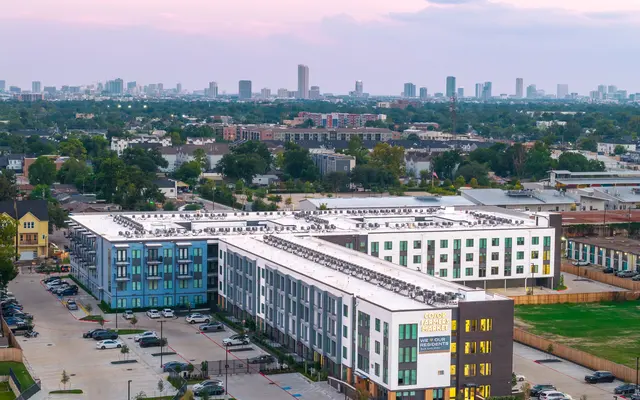 Aerial view of a modern apartment complex featuring multicolored buildings and ample parking spaces, with a city skyline in the background during sunset.