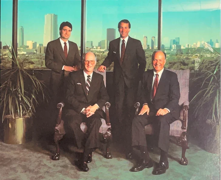 Group of Business Executives in Office A group of four men in formal suits posing for a photograph in a high-rise office with a city skyline visible through large windows. Two men are seated, and two are standing. There are decorative plants in the foreground.