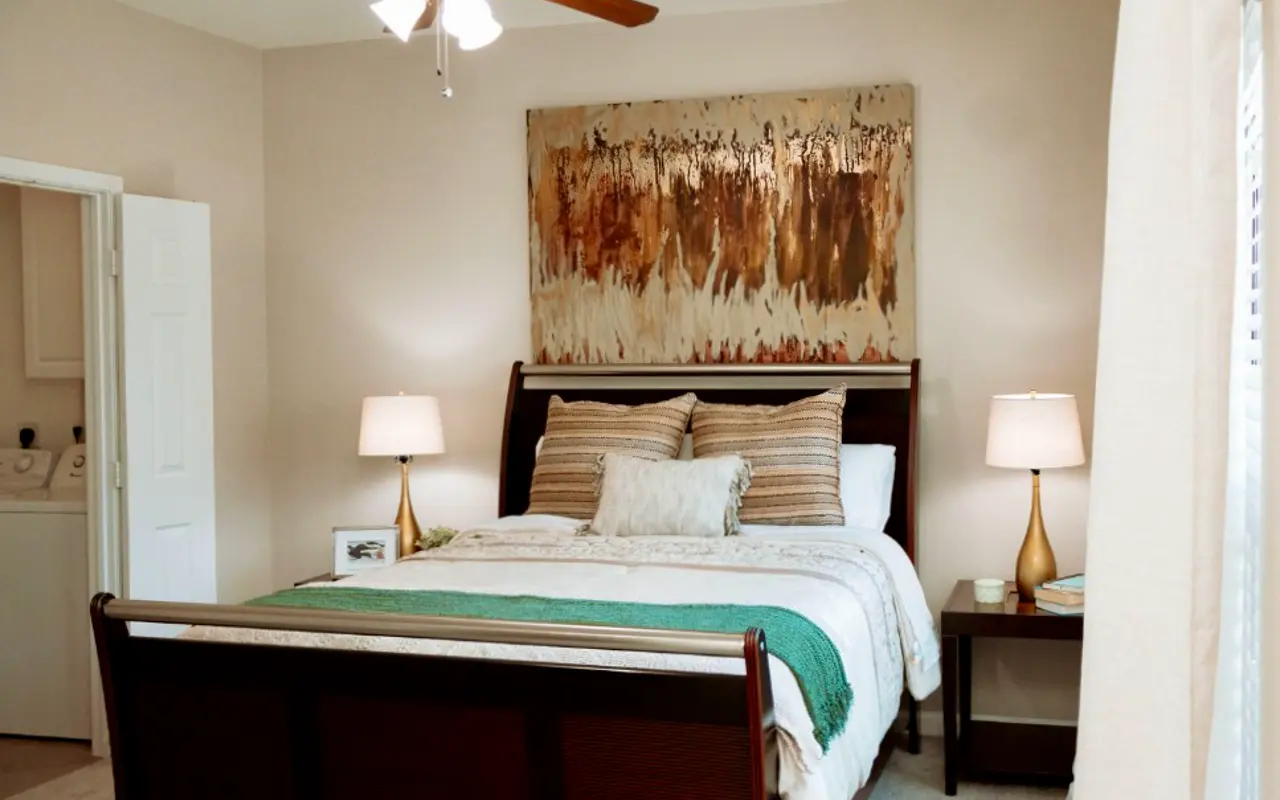 Cozy Bedroom Interior A cozy bedroom featuring a wooden bed frame with an assortment of pillows, a bedspread with green accents, nightstands with lamps, and a large piece of abstract art on the wall. A door leads to a laundry area on the left side.