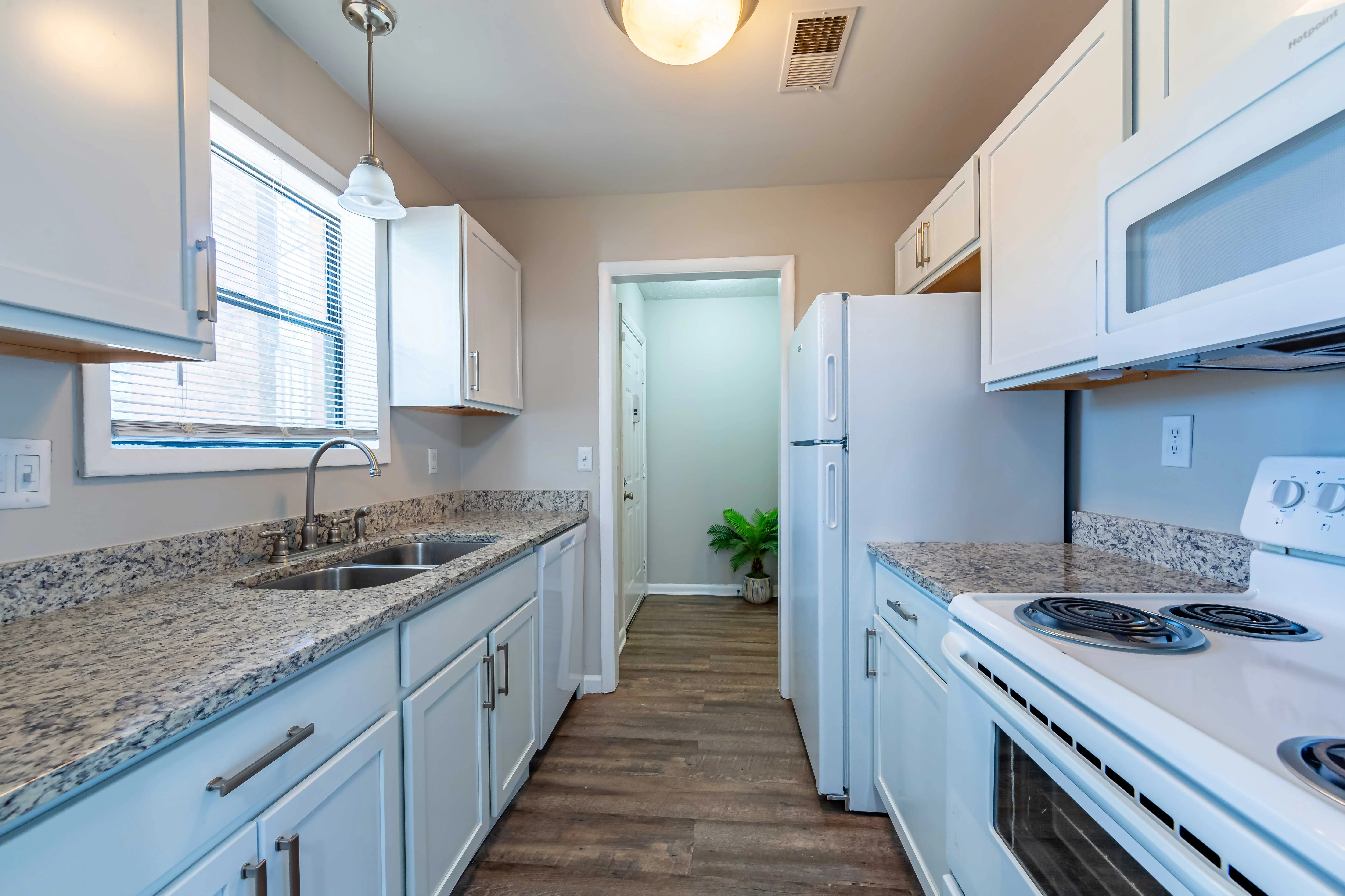 A modern kitchen featuring white cabinets, a countertop made of gray speckled granite, a double sink, and a white stove. There's a window allowing natural light and a door leading to a corridor. Stainless steel refrigerator is also visible.