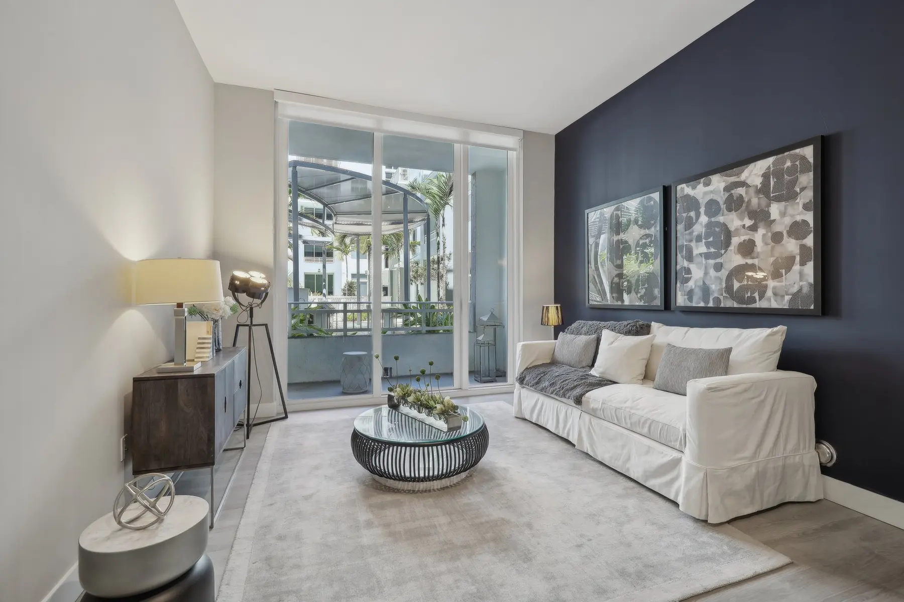 A stylish modern living room featuring a white sofa, a round coffee table, and large windows opening to a patio. The wall is painted in dark blue and there are two large abstract artworks on the wall.